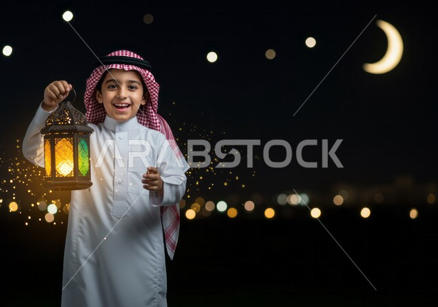 Celebrating the arrival of the month of goodness, decorations and lights for holidays and occasions, preparing and getting ready to receive the holy month of Ramadan, a smiling Emirati Gulf Arab boy wearing the traditional dress and shemagh holding a decorated illuminated metal lantern in his hand, looking at the camera with gestures of happiness and pleasure