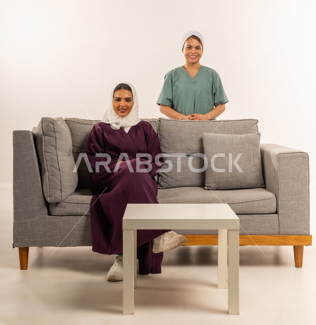 النظر الى الكاميرا بإيماءات السعادة والسرور، مدبرة منزل تستعين بخادمة أجنبية، استقدام العماله المنزليه المرخصه والمؤهله، توفير خدمات العناية بالمنزل، بورتريه لامرأة عربية خليجية سعودية محجبة ترتدي عباءة خمرية تجلس على اريكة وتقف خلفها عاملة فلبينية، خلفية بيضاء