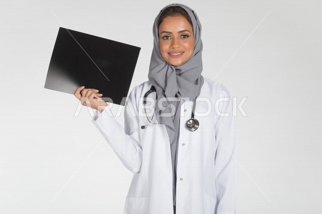 بورتريه لطبيبة عربية خليجية سعودية تمسك ورقة خاصة بالفحص الاشعاعي (الاشعة)، مفهوم الطب والرعاية الصحية، ايماءات وجه مبتسمة، تقديم الخدمات الصحية، خلفية بيضاء
