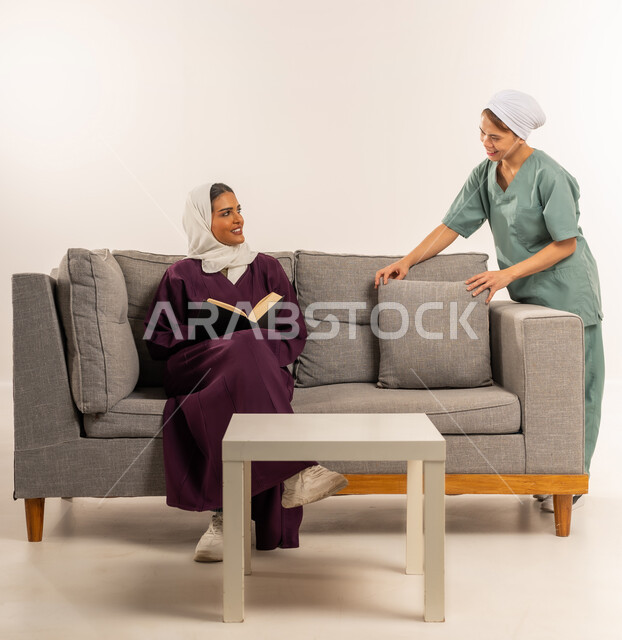 توجيه وإشراف مدبرة المنزل على خدمة التنظيف، عاملة نظافة تقوم بترتيب الوسائد، المهام والأعمال المنزلية الروتينية، بورتريه لسيدة عربية خليجية سعودية محجبة ترتدي عباءة خمرية تجلس على الأريكة تقوم بقراءة الكتب و تستعين بخادمة أجنبية داخل المنزل، خلفية بيضاء