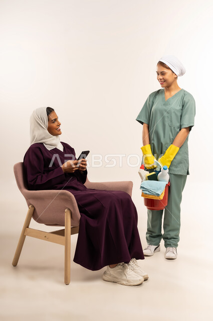 خادمات منزلية مدربة ومؤهلة، بورتريه لعاملة نظافة أجنبية تحمل بيدها سلة قش بأدوات تنظيف المنزل تتلقى تعليمات النظافة من ربة المنزل، سيدة عربية خليجية سعودية محجبة ترتدي عباءة خمرية تجلس على الأريكة تحمل الهاتف المحمول وترشد الخادمة الفلبينية، خلفية بيضاء