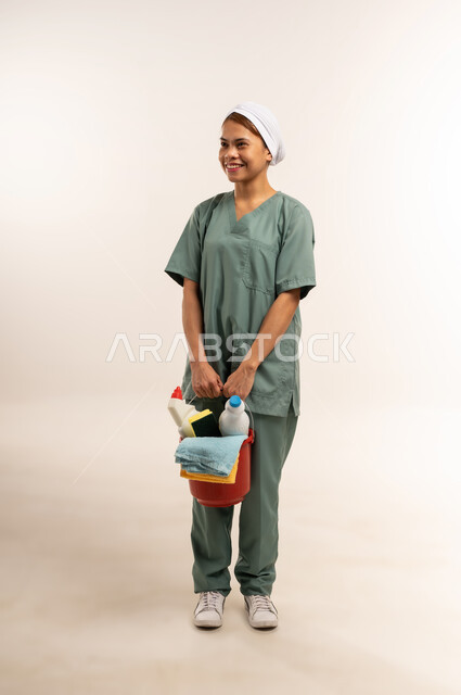 جودة الخدمات في السعودية، المهام والأعمال المنزلية الروتينية، وظائف ومهن نسائية، استقدام العمالة الاجنبية المدربة والمؤهلة، بورتريه لعاملة نظافة فلبينية ترتدي اليونيفورم الخاص بالعمل تنظر الى شيء ما وتحمل أدوات التنظيف، الطول الكامل للجسم، خلفية بيضاء