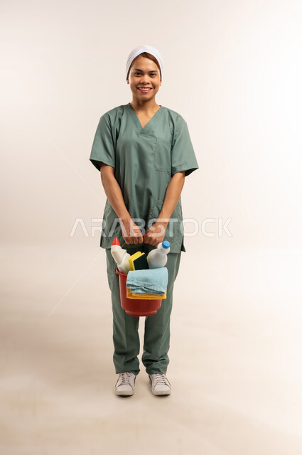 المهام والأعمال المنزلية الروتينية، جودة الخدمات في السعودية، وظائف ومهن نسائية، استقدام العمالة الاجنبية المدربة والمؤهلة، بورتريه لعاملة نظافة فلبينية ترتدي اليونيفورم الخاص بالعمل تنظر للكاميرا وتحمل أدوات التنظيف، الطول الكامل للجسم، خلفية بيضاء