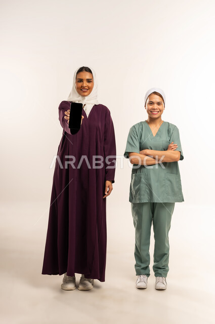 منصة وتطبيق لاستقدام العماله المنزليه المدربة، عرض شاشة سوداء فارغة عبر الهاتف المحمول، الخدمات الإلكترونيه المتقدمه لتوفير العاملات المؤهله، بورتريه لسيدة محجبة محجبة ترتدي عباءة خمرية تقف مع عاملة نظافة فلبينية ترتدي اليونيفورم، الطول الكامل للجسم، خلفية بيضاء