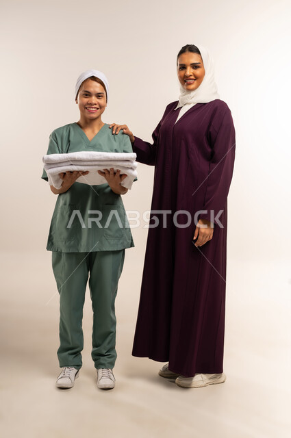 رضا ربة المنزل عن اداء العاملة، تأجير العمالة الخارجية المرخصة والمؤهلة، توفير خدمات العناية بالمنزل، المعاملة الحسنة للخادمات، بورتريه لسيدة عربية خليجية سعودية محجبة ترتدي عباءة خمرية تعطي الارشادات لخادمة تقوم بحمل الملابس المطوية، الطول الكامل للجسم، خلفية بيضاء
