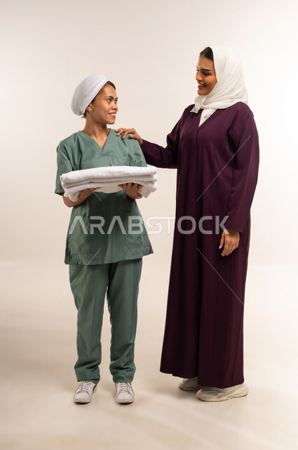 تأجير العمالة الخارجية المرخصة والمؤهلة، رضا ربة المنزل عن اداء العاملة، توفير خدمات العناية بالمنزل، المعاملة الحسنة للخادمات، بورتريه لسيدة عربية خليجية سعودية محجبة ترتدي عباءة خمرية تعطي الارشادات لخادمة تقوم بحمل الملابس المطوية، الطول الكامل للجسم، خلفية بيضاء