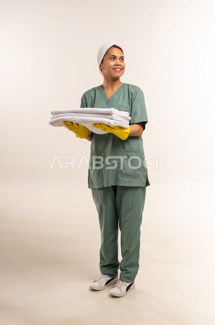 الأعمال والمهام المنزلية الروتينية، جودة الخدمات في السعودية، استقدام العمالة الاجنبية المدربة والمؤهلة، وظائف ومهن نسائية، بورتريه لعاملة نظافة فلبينية ترتدي اليونيفورم الخاص بالعمل والقفازات تحمل بيدها الثياب النظيفة المكوية، الطول الكامل للجسم، خلفية بيضاء