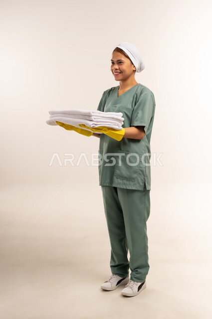 جودة الخدمات في السعودية، استقدام العمالة الاجنبية المدربة والمؤهلة، وظائف ومهن نسائية، الأعمال والمهام المنزلية الروتينية، بورتريه لعاملة نظافة فلبينية ترتدي اليونيفورم الخاص بالعمل والقفازات تحمل بيدها الثياب النظيفة المكوية، الطول الكامل للجسم، خلفية بيضاء