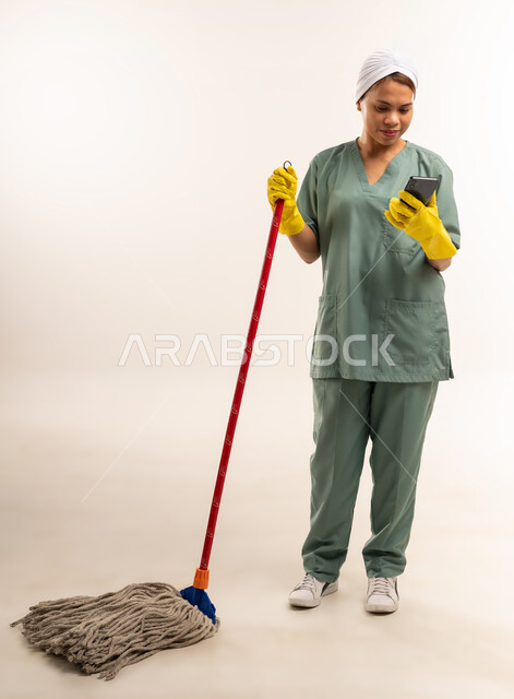 Quality of services in Saudi Arabia, routine household chores and tasks, recruitment of trained and qualified foreign workers, portrait of a Filipino cleaning lady wearing a work uniform and gloves holding a hand mop and looking at a mobile phone, full body length, white background