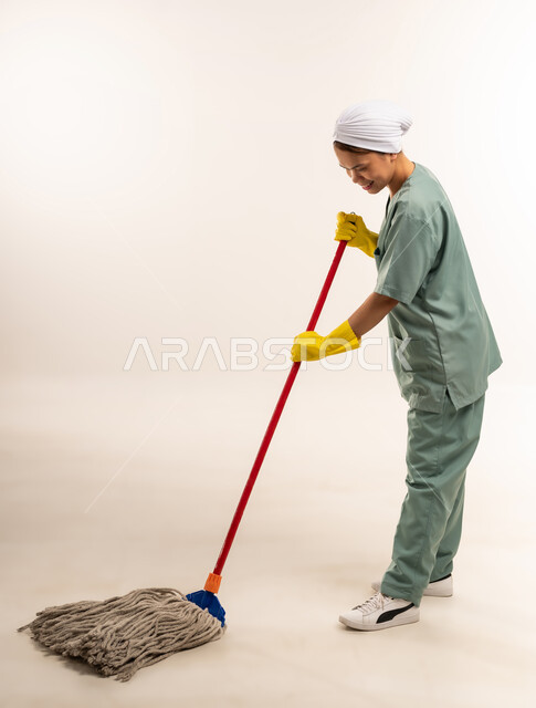 الأعمال والمهام المنزلية الروتينية، استقدام العمالة الاجنبية المدربة والمؤهلة، مهن ووظائف نسائية، جودة الخدمات في السعودية، بورتريه لعاملة نظافة فلبينية ترتدي اليونيفورم الخاص بالعمل والقفازات تقوم بتنظيف الأرض بالممسحة اليدوية، الطول الكامل للجسم، خلفية بيضاء