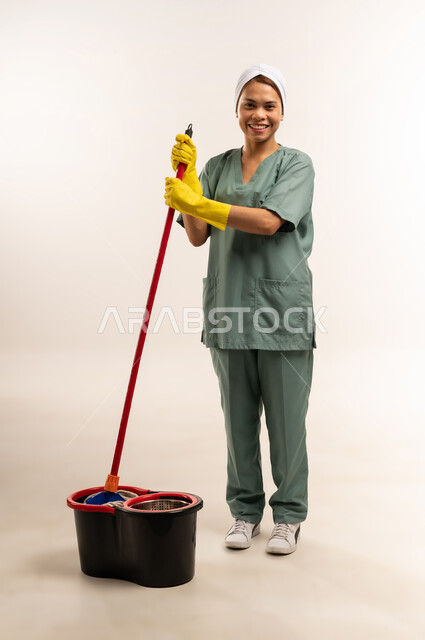 المهام والأعمال المنزلية الروتينية، استقدام العمالة الاجنبية المدربة والمؤهلة، مهن ووظائف نسائية، جودة الخدمات في السعودية، بورتريه لعاملة نظافة فلبينية ترتدي اليونيفورم الخاص بالعمل والقفازات تمسك الممسحة اليدوية للبدء بالتنظيف، الطول الكامل للجسم، خلفية بيضاء