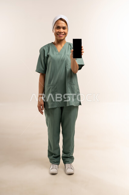 الخدمات الإلكترونيه المتقدمه لتوفير الخادمات والعاملات المؤهله، منصات إلكترونية لاستقدام العماله المنزليه المدربة، بورتريه لعاملة نظافة فلبينية مبتسمة ترتدي يونيفورم العمل وتحمل هاتف بشاشة سوداء فارغه، مهن ووظائف نسائية، الطول الكامل للجسم، خلفية بيضاء