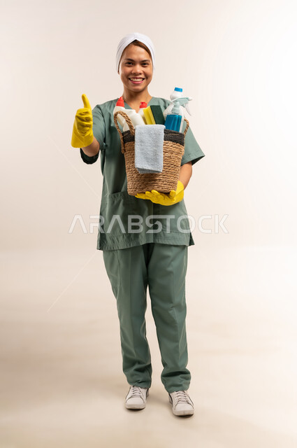 استقدام العمالة الاجنبية المدربة، الأعمال والمهام المنزلية الروتينية، جودة الخدمات في السعودية، بورتريه لعاملة نظافة فلبينية ترتدي اليونيفورم الخاص بالعمل والقفازات تحمل أدوات التنظيف وترفع اصبع الابهام بإيماءات الموافقة والاعجاب، الطول الكامل للجسم، خلفية بيضاء