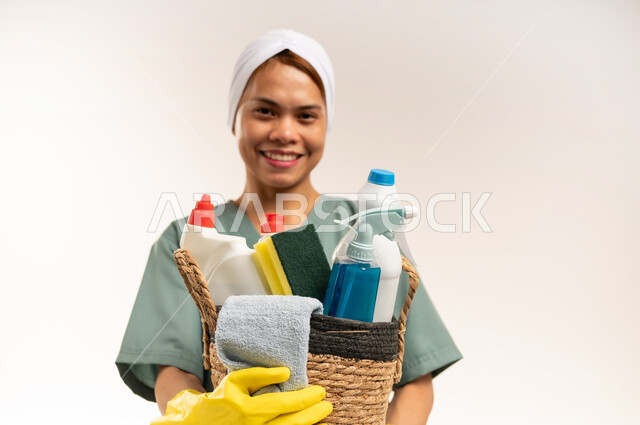 Women's jobs and professions, recruitment of trained foreign workers, routine household chores and tasks, quality of services in Saudi Arabia, close-up portrait of a Filipino cleaning lady wearing a work uniform and gloves looking at the camera and holding cleaning tools, white background
