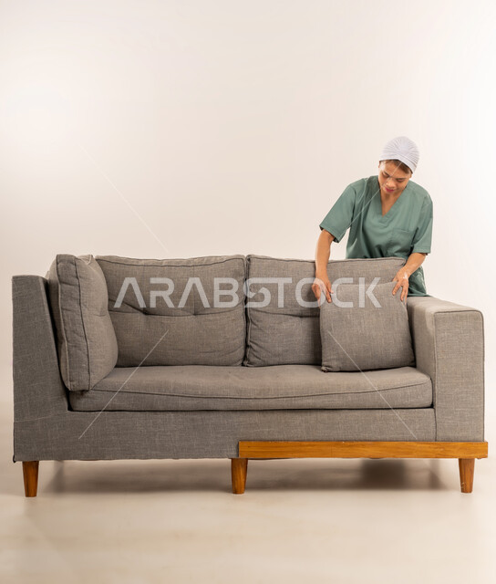 وظائف ومهن نسائية، استقدام العمالة الاجنبية المدربة والمؤهلة، الأعمال والمهام المنزلية الروتينية، بورتريه لعاملة نظافة فلبينية ترتدي اليونيفورم الخاص بالعمل وتقوم بترتيب الأريكة الرمادية، جودة الخدمات في السعودية، خلفية بيضاء