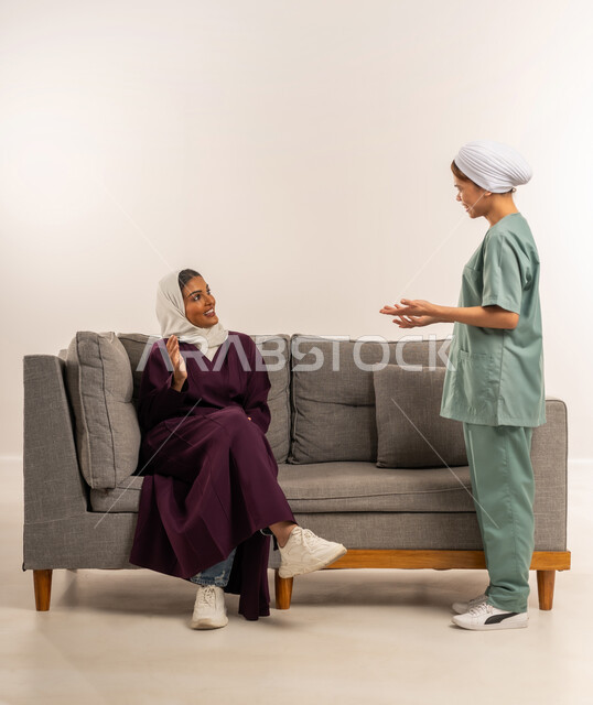 مدبرة منزل تستعين بخادمة أجنبية، استقدام العماله المنزليه المرخصه والمؤهله، توفير خدمات العناية بالمنزل، الإحسان في التعامل مع الخادمات في البيوت، بورتريه لامرأة عربية خليجية سعودية محجبة ترتدي عباءة خمرية تجلس على اريكة تتحدث مع العاملة الفلبينية، خلفية بيضاء