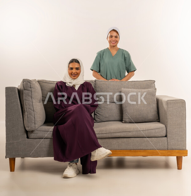 النظر الى الكاميرا بإيماءات السعادة والسرور، مدبرة منزل تستعين بخادمة أجنبية، استقدام العماله المنزليه المرخصه والمؤهله، توفير خدمات العناية بالمنزل، بورتريه لامرأة عربية خليجية سعودية محجبة ترتدي عباءة خمرية تجلس على اريكة وتقف خلفها عاملة فلبينية، خلفية بيضاء
