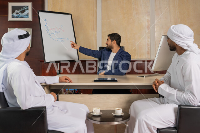 شرح خطط و استراتيجيات العمل و المعلومات المالية, مفهوم الاستثمار الاقتصادي, رجل أعمال عربي مبتسم يرتدي الزي الرسمي يجلس في المكتب داخل مقر الشركة يشير الى بورد ابيض, سيدان عربيان خليجيان اماراتيان يرتديان الزي التقليدي يقومان بالاتفاق على انجاز المهام