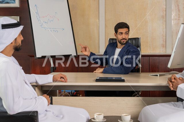 Discussion of economics and financial information, a smiling Arab businessman in formal dress sitting in the office inside the company headquarters pointing to a white board, the concept of economic investment, an Arab Gulf Emirati gentleman wearing a ghutra and kandura agreeing to complete tasks