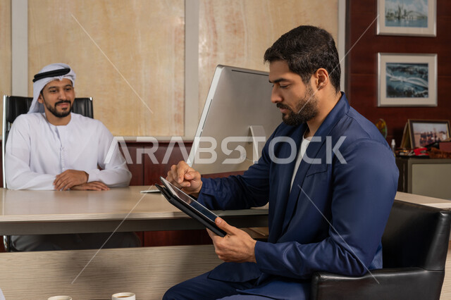 الاطلاع على كيفية العمل باستخدام جهاز التابليت، تحقيق اهداف الشركة، مكاتب و شركات خليجية، الوحدة والعمل الجماعي لتطوير العمل وحل المشكلات، رجل أعمال عربي خليجي اماراتي يرتدي الغترة و الكندورة يجلس مع الموظف في المكتب, مهنة ووظيفة اماراتية