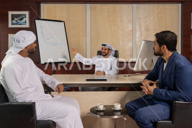العمل الجماعي وروح الفريق، مناقشة وضمان فهم واضح وشامل للاعمال، استعمال السبورة لتوضيح العمليات والاجراءات المتبعة في الشركة، رجل عربي اماراتي خليجي يرتدي الزي التقليدي يجلس في مكتبه مع زملائه يقوم بشرح المخططات، الاحترافية والتفاني في الإنجاز