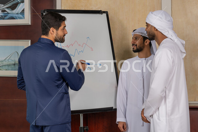 مناقشة وضمان فهم واضح وشامل للاعمال، رجلان عربيان اماراتيان خليجيان يرتديان الكندورة يقفان مع رجل بالبدلة الرسمية يستعمل السبورة لتوضيح العمليات والاجراءات المتبعة في الشركة، العمل الجماعي وروح الفريق