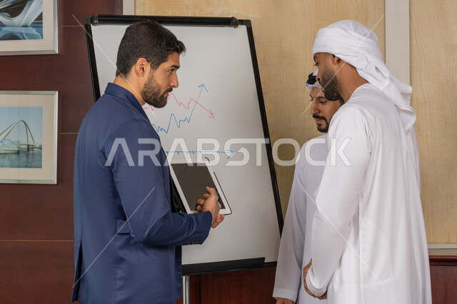 Teamwork and team spirit, using the whiteboard to explain the company’s processes and procedures, discussing and ensuring a clear and comprehensive understanding of the business, two Emirati Gulf Arab men wearing the kandura with a man in a formal suit using a portable tablet to explain, professionalism and dedication to achievement
