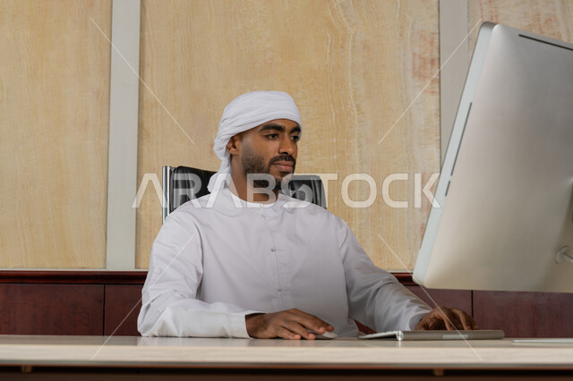 دراسه المشروع ومراجعه التقرير السنوي، استخدام التقنيات الحديثة المتطورة، جلسه عمل عن بعد, أداء المهمة اليومية داخل مقر الشركه، رجل أعمال عربي خليجي اماراتي يرتدي الكندورة العمامة يقوم بالعمل على جهاز الحاسوب المكتبي، دمج التقنية والتكنولوجيا في الحياة العملية