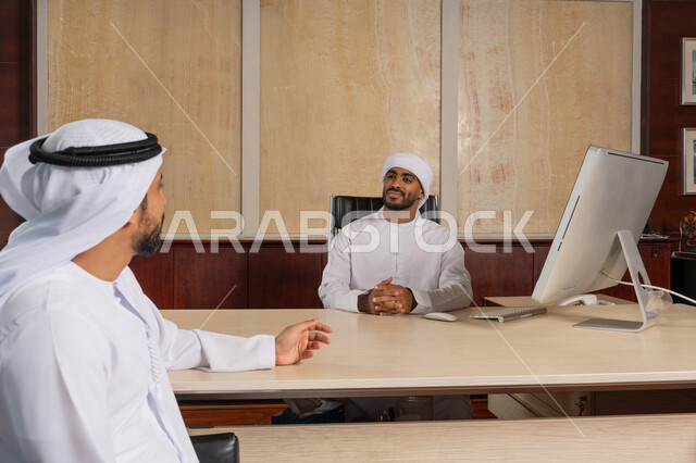 تبادل النقاشات في مهام العمل, مهنة ووظيفة اماراتية, شرح خطط و استراتيجيات المهام المطلوبة, التعارف وبناء العلاقات الاجتماعية, رجلان أعمال عربيان خليجيان اماراتيان يرتدي الكندورة يجلسان في المكتب داخل مقر الشركة، الشراكة في المشاريع والأعمال