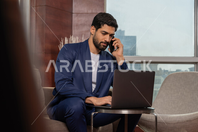 Remote working session, using advanced modern technologies, making a voice call via mobile phone, studying the project and reviewing the annual report, an Arab Gulf Emirati businessman wearing a formal uniform working on a computer, integrating technology and technology into practical life