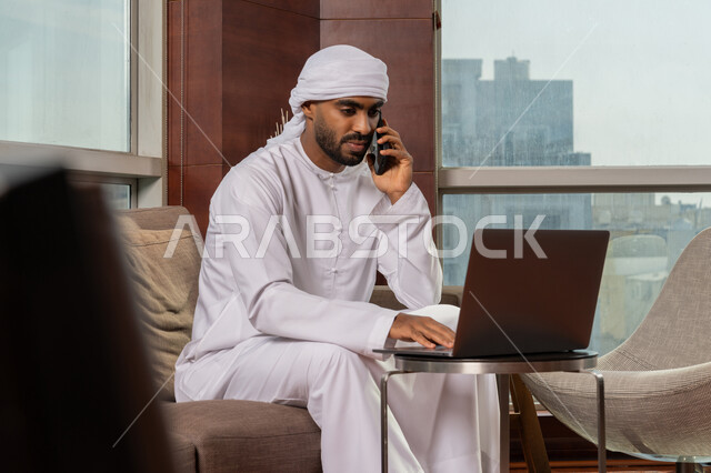 اجراء مكالمة هاتفية عبر الجوال, أداء المهام اليومية داخل مقر الشركه، رجل عربي خليجي اماراتي يرتدي الكندوره و العمامة البيضاء يقوم بالعمل على جهاز الحاسوب المحمول، دراسه المشاريع ومراجعه التقارير السنويه، جلسه عمل عن بعد, استخدام التقنية الحديثة المتطورة