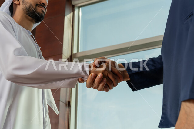 الشراكة في الأعمال والمشاريع، صداقات وتعارف وبناء علاقات اجتماعية، عقد صفقات عمل جديدة ناجحة، صورة مقربة ليد سيد عربي خليجي اماراتي يرتدي الكندورة يقوم بمصافحة رجل أعمال داخل مقر الشركة، توسيع مجالات العمل