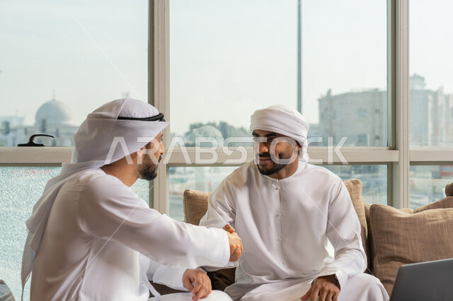 الوحدة والتفاعل الجماعي, الاتفاق لتوقيع صفقة شراكة جديدة، مفهوم الإدارة المتعاونة، رجلان عربيان خليجيان اماراتيان يرتديان الكندوة يجلسان في مقر الشركة يصافحان بعضهما بإيماءات السرور، تحقيق الأهداف وحل مشكلات العمل باستخدام التقنية الحديثة المتطورة