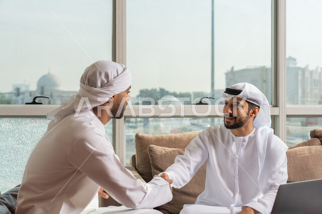 الاتفاق لتوقيع صفقة شراكة جديدة، الوحدة والتفاعل الجماعي, مفهوم الإدارة المتعاونة، رجلان عربيان خليجيان اماراتيان يرتديان الكندوة يجلسان في مقر الشركة يصافحان بعضهما بإيماءات السرور، تحقيق الأهداف وحل مشكلات العمل باستخدام التقنية الحديثة المتطورة