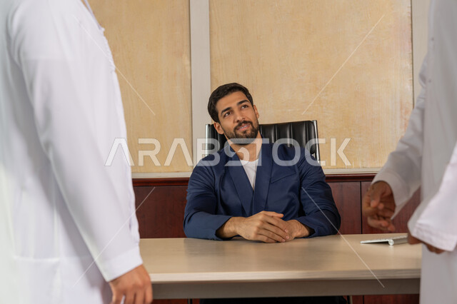 Exchanging discussions on work tasks, Emirati profession and job, comfortable environment in Emirati companies, explaining plans and strategies for required tasks, getting to know each other and building social relationships, an Arab Gulf Emirati businessman wearing a formal uniform sitting in the office inside the company headquarters looking at one of the employees