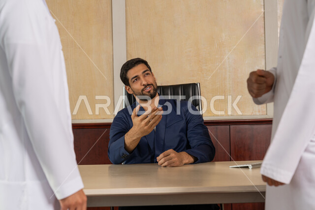 Comfortable environment in UAE companies, explaining work plans and strategies, getting to know each other and building social relationships, an Emirati Gulf Arab businessman wearing a formal uniform sitting in the office inside the company headquarters, pointing to one of the employees, an Emirati profession and job