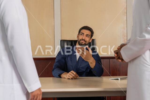 شرح خطط و استراتيجيات العمل, التعارف وبناء العلاقات الاجتماعية, رجل أعمال عربي خليجي اماراتي يرتدي الزي الرسمي يجلس في المكتب داخل مقر الشركة يقوم بالاشارة الى أحد الموظفين, مهنة ووظيفة اماراتية, بيئة مريحة في الشركات الاماراتية