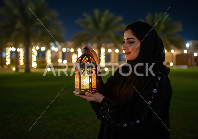  الاستعداد والتجهز لاستقبال شهر رمضان المبارك، صورة مقربة من الجانب لإمرأة عربية خليجية سعودية محجبة تحمل فانوس معدني مزخرف مضيء تقف في الهواء الطلق، زينة وأنوار الأعياد والمناسبات الدينية الاسلامية، الاحتفال بقدوم شهر الخير في احدى الحدائق العامة
