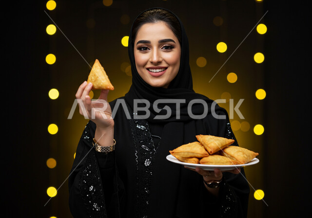 فطائر رمضانية شهية، النظر الى الكاميرا بايماءات السعادة والسرور بقدوم شهر الخير، طبق السمبوسه اللذيذ، صورة مقربة لإمرأة عربية خليجية سعودية مبتسمة ترتدي الحجاب والعباءة تمسك طبق من المقبلات في شهر رمضان المبارك، خلفية سوداء