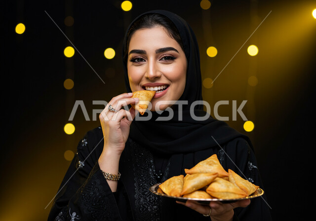طبق السمبوسه اللذيذ، فطائر رمضانية شهية، النظر الى الكاميرا بايماءات السعادة والسرور بقدوم شهر الخير، صورة مقربة لإمرأة عربية خليجية سعودية مبتسمة ترتدي الحجاب والعباءة تمسك طبق من المقبلات في شهر رمضان المبارك، خلفية سوداء