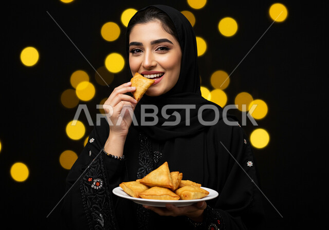 فطائر رمضانية شهية، النظر الى الكاميرا بايماءات السعادة والسرور بقدوم شهر الخير، طبق السمبوسه اللذيذ، صورة مقربة لإمرأة عربية خليجية سعودية مبتسمة ترتدي الحجاب والعباءة تمسك طبق من المقبلات في شهر رمضان المبارك، خلفية سوداء