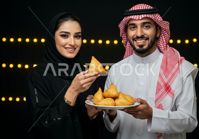 طبق السمبوسه اللذيذ، صورة مقربة لزوجين عربين خليجيين سعوديين مبتسمين يمسكان طبق من المقبلات في شهر رمضان المبارك، النظر الى الكاميرا بايماءات السعادة والسرور بقدوم شهر الخير، فطائر رمضانية شهية، خلفية سوداء
