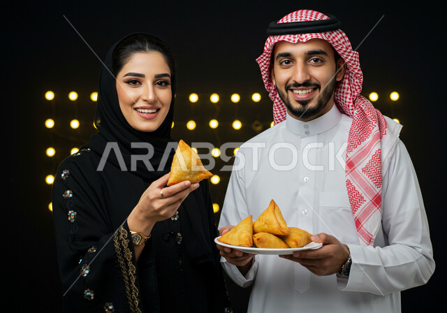 طبق السمبوسه اللذيذ، صورة مقربة لزوجين عربين خليجيين سعوديين مبتسمين يمسكان طبق من المقبلات في شهر رمضان المبارك، النظر الى الكاميرا بايماءات السعادة والسرور بقدوم شهر الخير، فطائر رمضانية شهية، خلفية سوداء