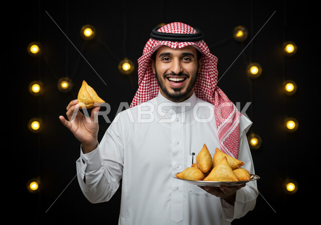 النظر الى الكاميرا بايماءات السعادة والسرور بقدوم شهر الخير، فطائر رمضانية شهية، طبق السمبوسه اللذيذ، صورة مقربة لرجل عربي خليجي سعودي مبتسم يرتدي الشماغ والثوب التقليدي يمسك طبق من المقبلات في شهر رمضان المبارك، خلفية سوداء