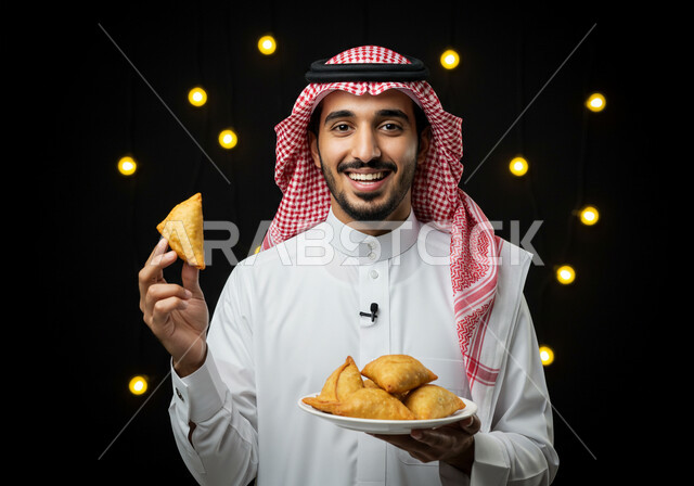 فطائر رمضانية شهية، النظر الى الكاميرا بايماءات السعادة والسرور بقدوم شهر الخير، طبق السمبوسه اللذيذ، صورة مقربة لرجل عربي خليجي سعودي مبتسم يرتدي الشماغ والثوب التقليدي يمسك طبق من المقبلات في شهر رمضان المبارك، خلفية سوداء