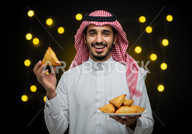 النظر الى الكاميرا بايماءات السعادة والسرور بقدوم شهر الخير، فطائر رمضانية شهية، طبق السمبوسه اللذيذ، صورة مقربة لرجل عربي خليجي سعودي مبتسم يرتدي الشماغ والثوب التقليدي يمسك طبق من المقبلات في شهر رمضان المبارك، خلفية سوداء