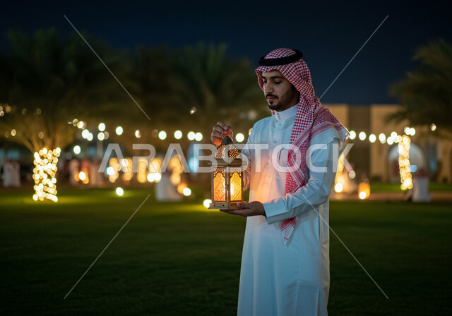 الاستعداد والتجهز لاستقبال شهر رمضان المبارك، الاحتفال بقدوم شهر الخير في احدى الحدائق العامة، صورة مقربة من الجانب لرجل عربي خليجي سعودي مبتسم يرتدي الشماغ والثوب التقليدي يمسك فانوس معدني مزخرف مضيء يقف في الهواء الطلق، زينة وأنوار الأعياد والمناسبات 