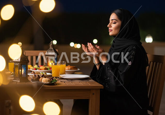 الجلوس على مائدة رمضان وشكر الله، الدعاء المستجاب وقت آذان المغرب، إمرأة عربية خليجية سعودية ترتدي الحجاب والعباءة ترفع يديها وتدعو الله تعالى في شهر الخير، الافطار في احد مطاعم المملكة العربية السعودية، سفرة طعام مليئة بأصناف من المأكولات والمقبلات الشهية