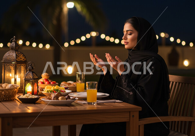 سفرة طعام مليئة بأصناف من المأكولات والمقبلات الشهية، الجلوس على مائدة رمضان وشكر الله، الدعاء المستجاب وقت آذان المغرب، إمرأة عربية خليجية سعودية ترتدي الحجاب والعباءة ترفع يديها وتدعو الله تعالى في شهر الخير، الافطار في احد مطاعم المملكة العربية السعودية