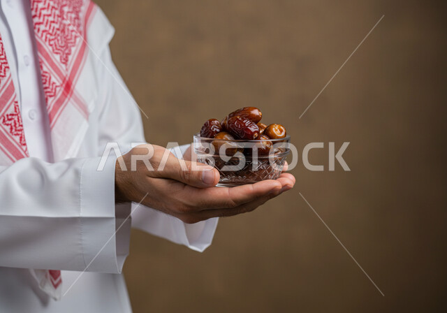 حسن الاستقبال وكرم الضيافة في شهر رمضان المبارك، وجبة الرطب الصحية ذات القيمة الغذائية العالية, محصول زراعي محلي وطني سعودي، بورتريه مقرب ليد رجل عربي خليجي سعودي يرتدي الثوب التقليدي والشماغ يحمل بيده وعاء مليء بحبات التمر الطازجة, خلفيه بنية